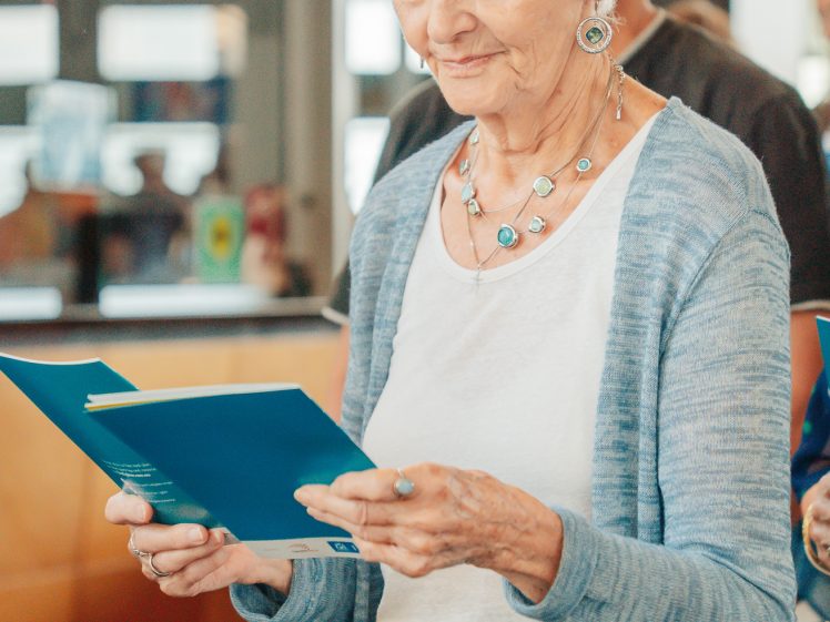 Image of An elderly woman with white hair, wearing a light blue cardigan and jewellery, stands indoors holding and reading two blue papers, appearing focused and content.