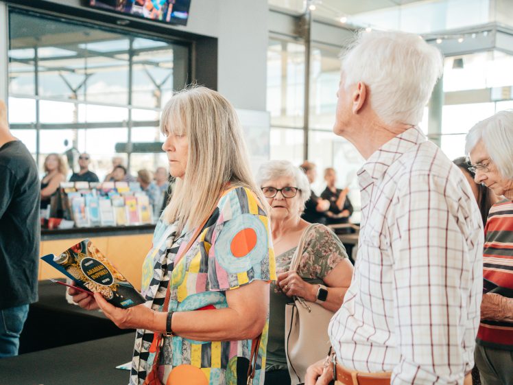 Image of A group of older adults stand indoors; a woman in a colourful top reads a leaflet while others queue. Sunlight streams through large windows in the background.