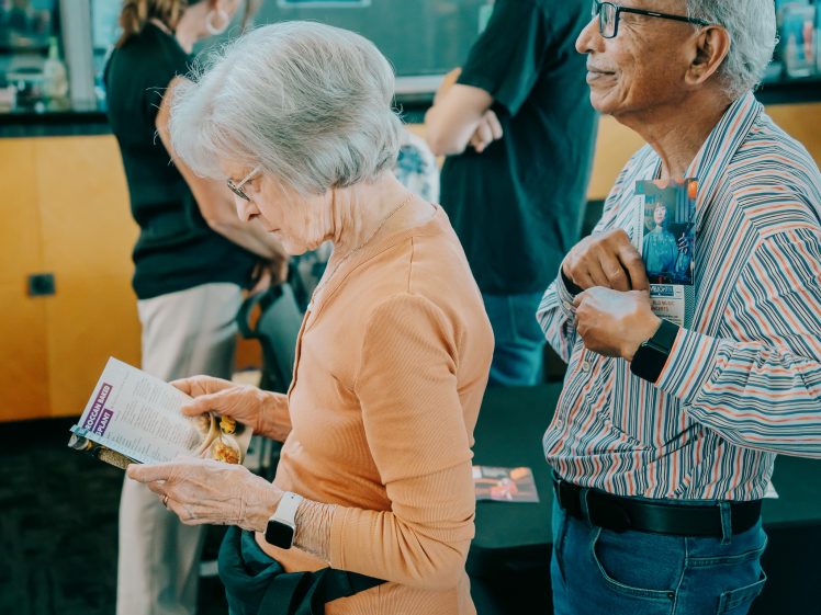 Image of Two elderly people stand in a queue; the woman in front reads a leaflet while the man behind her holds a flyer to his chest and smiles. Other people are seen blurred in the background.