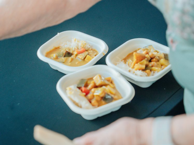 Image of Three bowls of rice topped with vegetable curry are placed on a dark surface. A person’s arm, partially visible, is serving or arranging the bowls.
