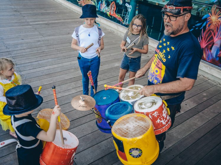 Image of A man playing colourful bucket drums leads a group of children who are tapping drumsticks on buckets. They stand on a wooden decking near a mural with vibrant, abstract designs.