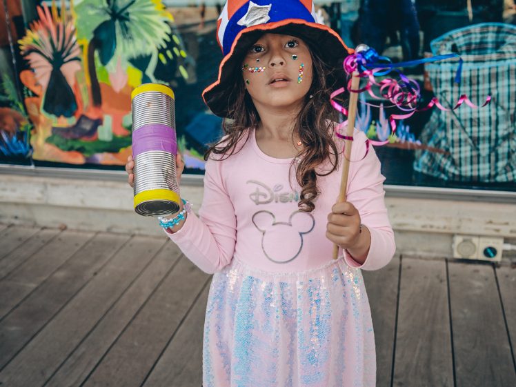 Image of A young girl in a sparkly dress, holding a decorated stick and a glittery container, wears a red, white, and blue hat and has face paint, standing on a wooden decking in front of a colourful mural.