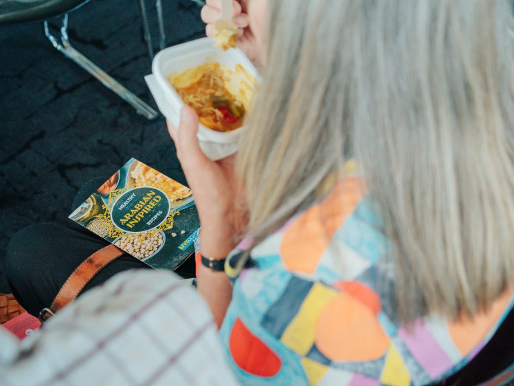 Image of A person with long, grey hair wearing a colourful, geometric-patterned shirt eats from a white container. An Asian Noodle Pot meal package is visible on their lap. The setting appears casual and informal.