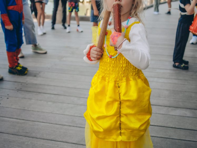Image of A young girl in a yellow princess dress holds two ice lollies and stands barefoot on a wooden decking at an outdoor event, with other people and children in fancy dress in the background.