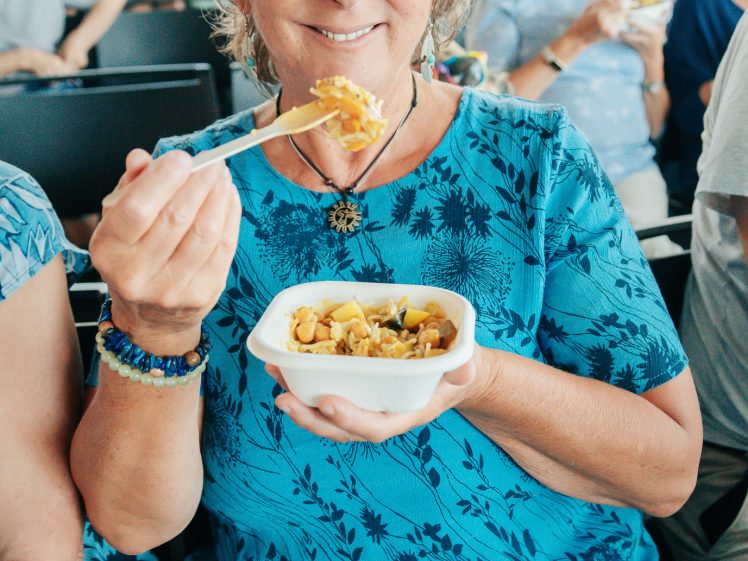 Image of An older woman in a blue dress smiles widely whilst holding a fork and a bowl of food at a crowded indoor event. Other people are seated and eating in the background.
