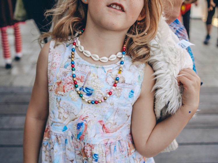 Image of A young girl with wavy hair wears a colourful beaded necklace, a shell necklace, and a dress with a map pattern. She has a jewel decoration on her forehead and stands outdoors, looking at the camera.