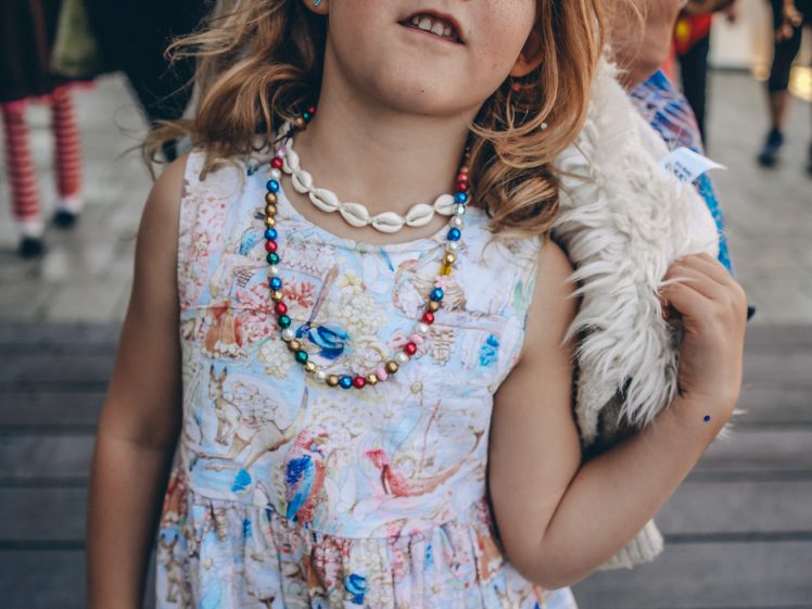 Image of Young girl with wavy hair wears a colourful dress, a shell necklace, and beaded jewellery. She has face paint on her forehead and stands outdoors, looking confidently at the camera. An adult’s arm rests gently on her shoulder.