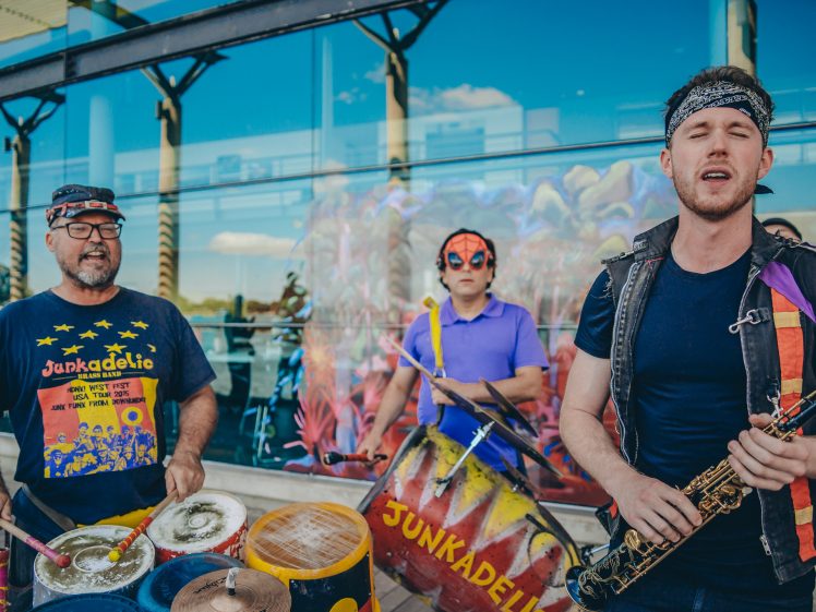 Image of Three musicians perform outdoors. Two play painted drums labelled Junkadelic, while one plays a saxophone. The drummer on the left wears glasses and a headband; the middle musician wears a bright mask and purple shirt.