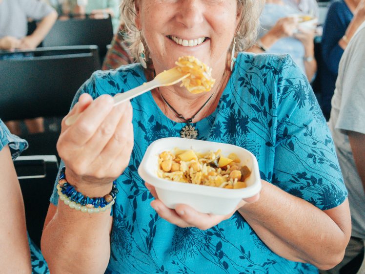 Image of A smiling older woman in a blue dress holds a takeaway container of food and a fork, surrounded by people in a bright, busy indoor setting.