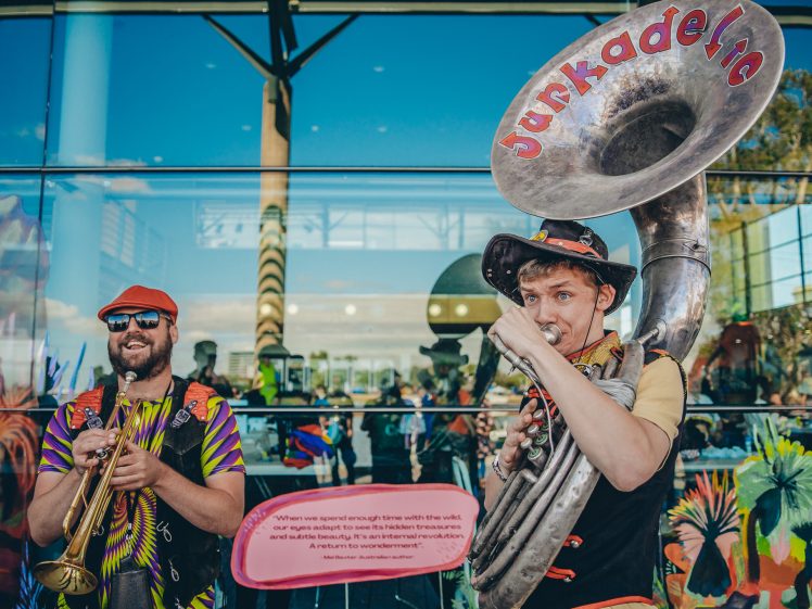 Image of Two musicians perform outside a glass building. One plays a trumpet and smiles, whilst the other plays a sousaphone labelled Junkadelic. Both wear colourful outfits and hats, with vivid reflections and artwork on the glass behind them.