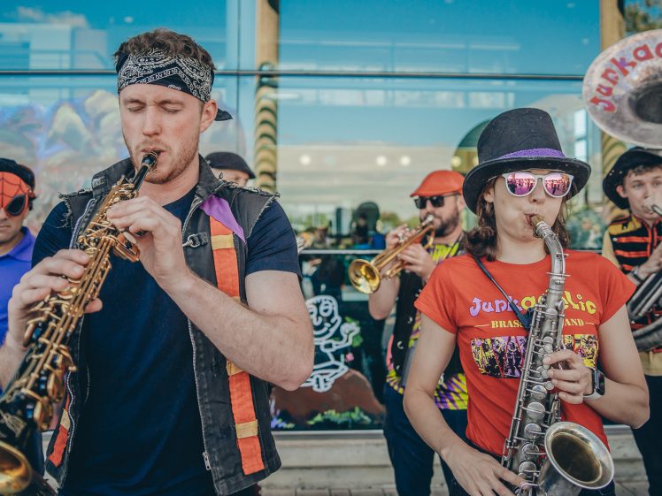 Image of A lively street band performs outdoors. Members play saxophones, trumpet, and sousaphone, wearing colourful clothes, hats, sunglasses, and fancy dress. A mural and glass building are visible in the background.