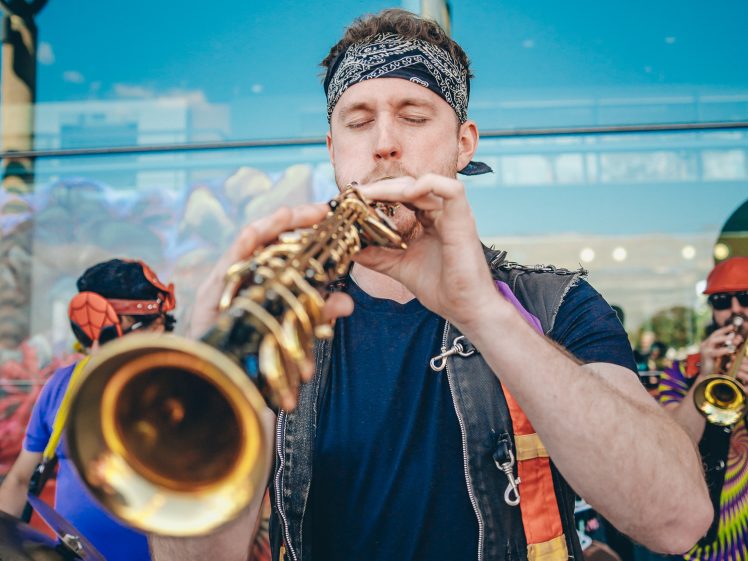 Image of A musician wearing a bandana and black waistcoat plays a brass instrument outdoors. Other performers in colourful clothing can be seen in the background, reflecting a lively street or festival setting.