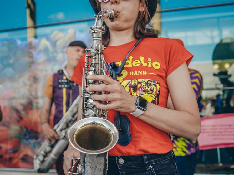 Image of A woman wearing a black hat, sunglasses, and a red “Psychedelic Band” shirt plays a saxophone outdoors. Another musician stands in the background. Reflections and colourful art appear on the glass wall behind them.