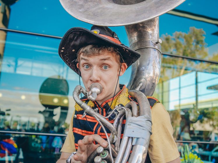Image of A young musician in a quirky hat plays a large silver sousaphone labelled Junkadelic outside a glass building, looking at the camera with a playful expression.