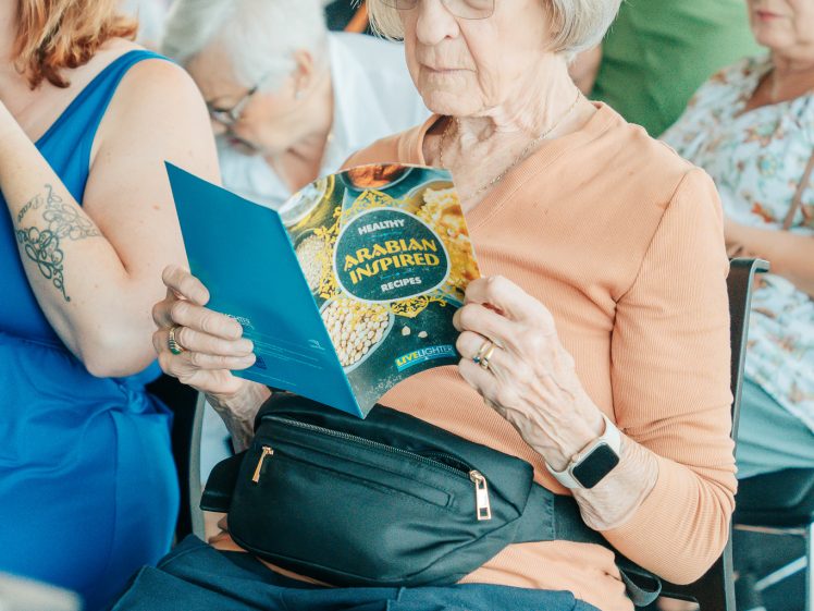 Image of An elderly woman with grey hair and glasses sits and reads a blue programme. She wears a peach top, bum bag, and smartwatch. Other older adults sit around her, some engaged in their own activities.