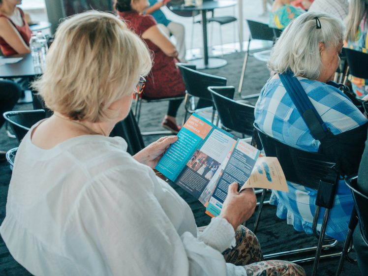Image of A woman with blonde hair and glasses sits reading a colourful brochure in a room with other seated people. The atmosphere appears casual and social, with round tables and bright daylight.