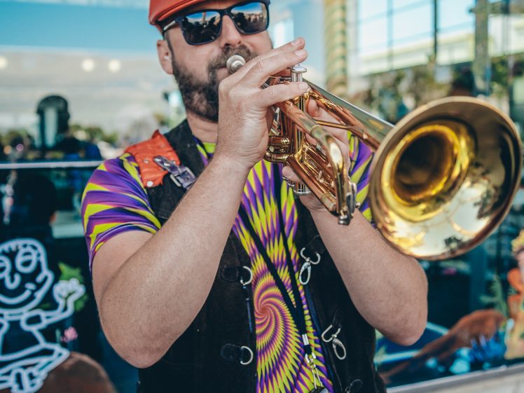 Image of A man wearing a red cap, sunglasses, and a colourful tie-dye shirt plays a brass trumpet outdoors. The background has glass windows with artistic drawings and reflections of trees and people.