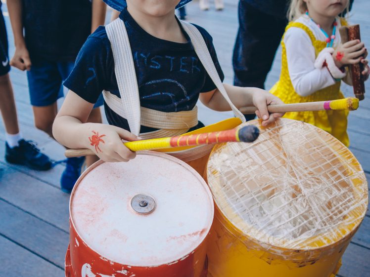 Image of A young boy wearing a blue hat plays makeshift drums made from large plastic buckets, using drumsticks, whilst other children stand nearby on a wooden floor.