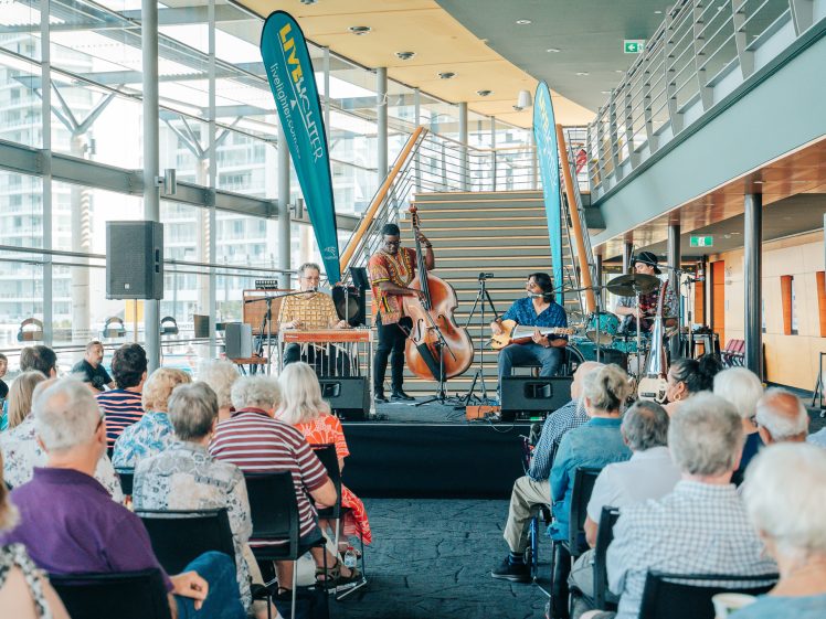 Image of A small band performs on a stage in front of an audience seated indoors. The band features a double bass, guitar, and keyboard, with large windows and a staircase in the background.