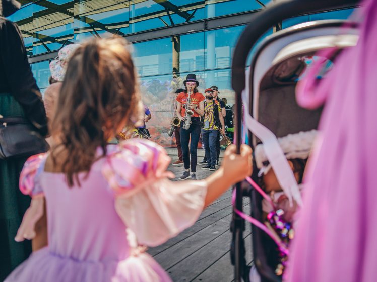 Image of A child in a pink princess dress stands near a pushchair, watching a group of musicians perform outdoors. The scene is lively, with people gathered around under bright daylight.