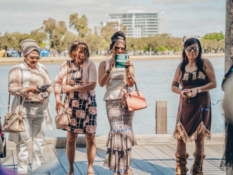 Image of Four women stand on a lakeside wooden decking, dressed in various stylish outfits, some holding mobiles or tablets. Trees and a modern building are visible in the background across the water.