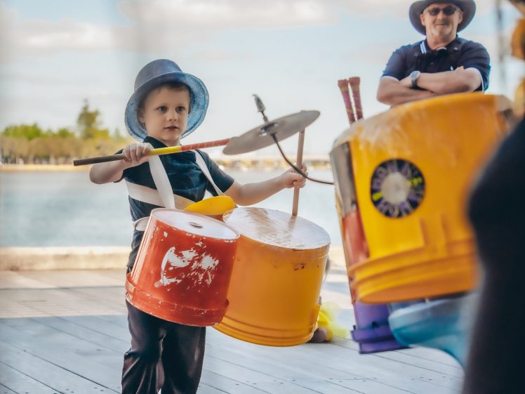 Image of A young boy wearing a hat plays a drum kit made from colourful recycled buckets outdoors, while an adult in a hat stands in the background, smiling and watching.