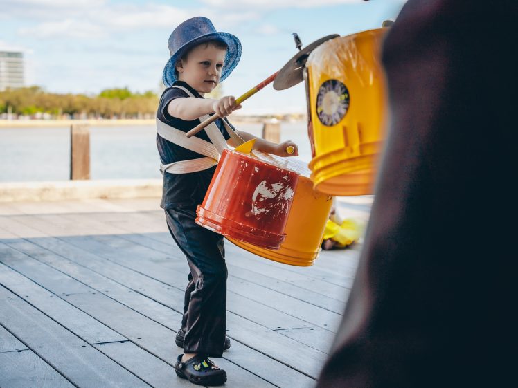 Image of A young child wearing a blue hat plays makeshift drums made from yellow and orange buckets, using drumsticks, outdoors near water on a wooden decking.