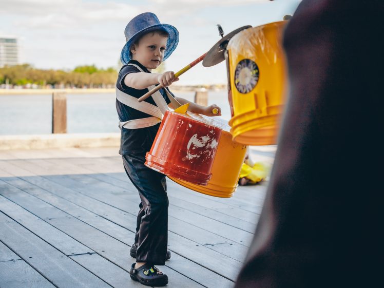 Image of A young child in a blue hat plays makeshift drums made from yellow and orange buckets outdoors near water, using drumsticks, with trees and buildings in the background.
