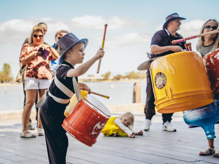Image of A young boy wearing a hat plays drums made from large colourful containers, surrounded by adults and children outdoors near water. People watch and take photos under a bright sky.