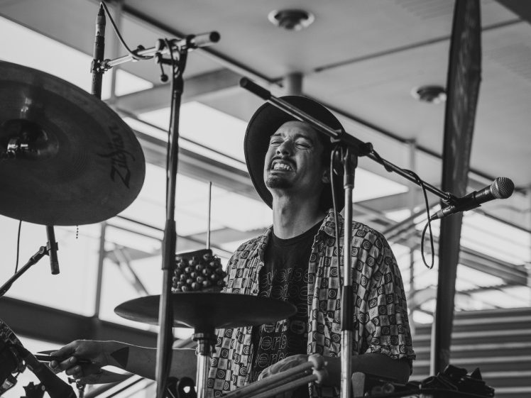 Image of A drummer wearing a hat and patterned shirt plays happily on stage, eyes closed and smiling, surrounded by drum equipment and microphones in a bright indoor setting.