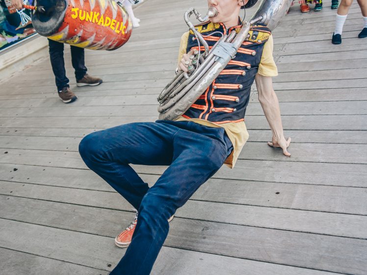 Image of A musician wearing a decorated hat and waistcoat plays a sousaphone while leaning back on a wooden boardwalk. Other band members in colourful costumes and instruments stand in the background.