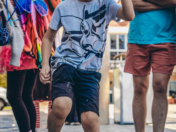 Image of A young boy in a cap and graphic T-shirt dances energetically outdoors, while a smiling man and a person in a colourful strawberry costume watch in the background.