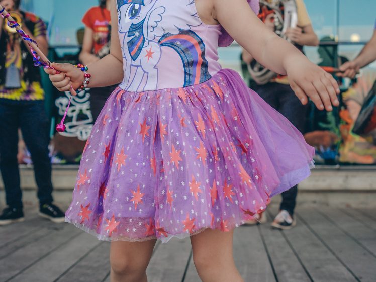 Image of A young child wearing a purple tutu dress with a unicorn design, striped socks, and black shoes poses confidently outdoors with a wand. A band plays in the background.