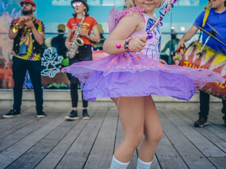 Image of A young child in a pink and purple tutu, holding a wand and wearing striped socks, dances on a wooden decking. Behind them, a band performs with instruments in colourful outfits.