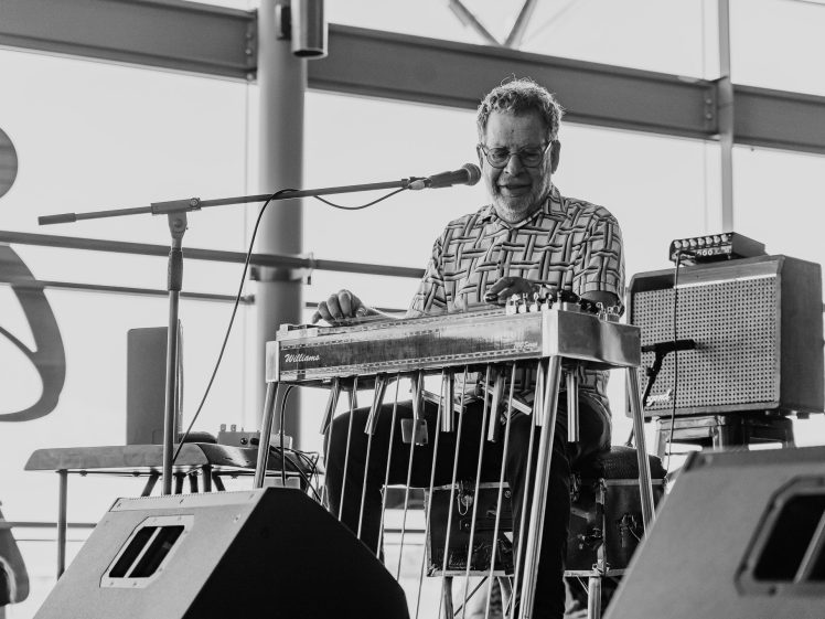Image of A man wearing sunglasses plays a pedal steel guitar and sings into a microphone on stage, with amplifiers beside him. The scene is indoors with large windows in the background. The image is in black and white.