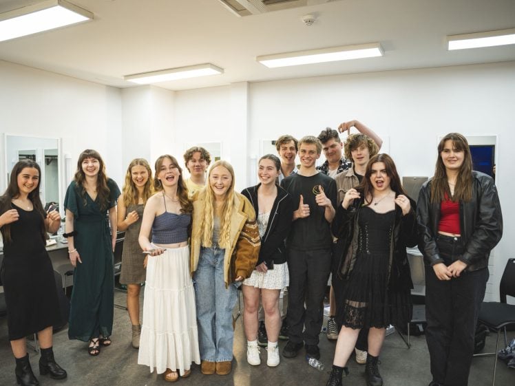 Image of A group of teenagers stand together in a brightly lit room, smiling and posing playfully for the camera. Some wear casual outfits, others are dressed up, and the atmosphere is cheerful and friendly.