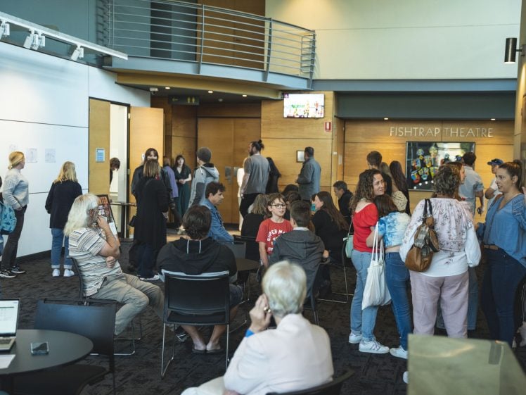 Image of A group of people gathers and chats in the lobby outside Fishtrap Theatre. Some are seated at tables, whilst others stand or walk around. The atmosphere is lively and sociable, with natural light from windows.