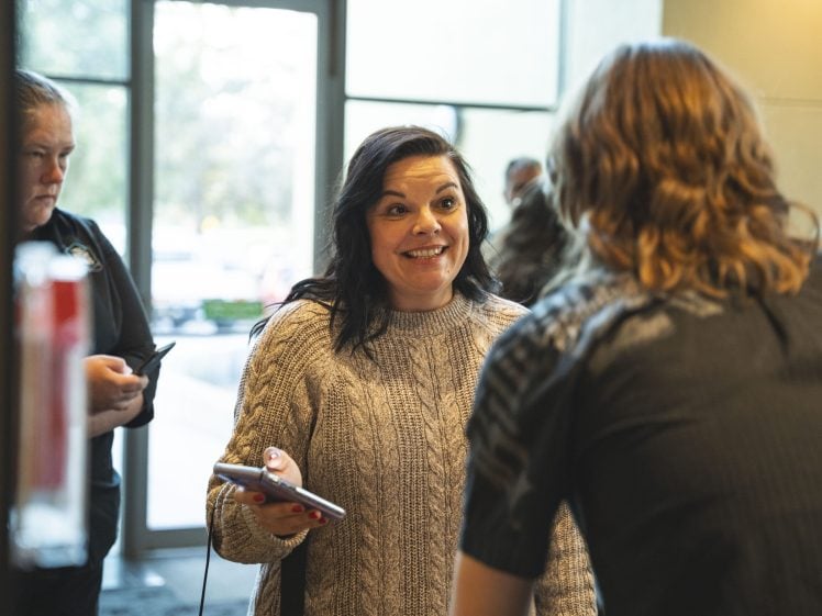 Image of A woman in a grey jumper smiles enthusiastically whilst holding her mobile and talking to another person indoors. Other people are visible in the background near a large window.
