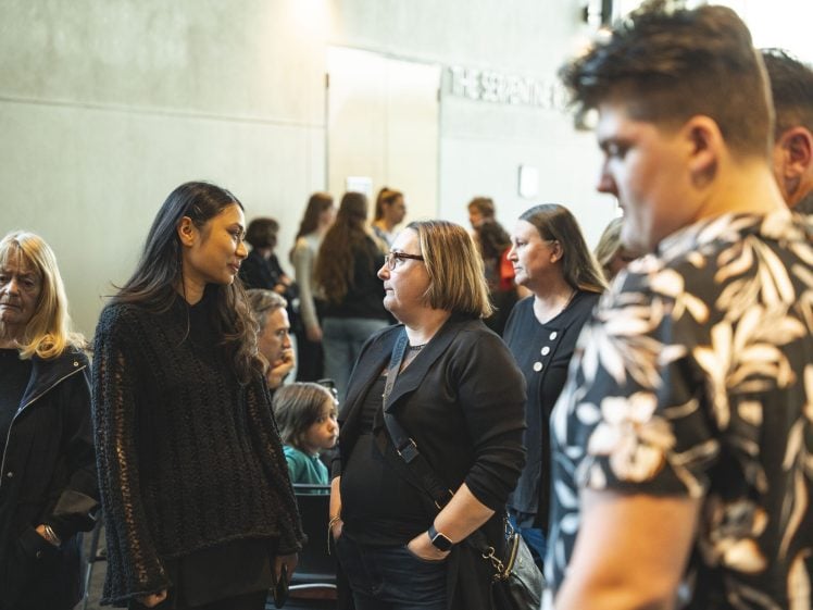 Image of A group of people stand indoors, engaged in conversations. Some are talking in pairs whilst others look around. The background shows more people, including a child seated, in a bright and modern setting.