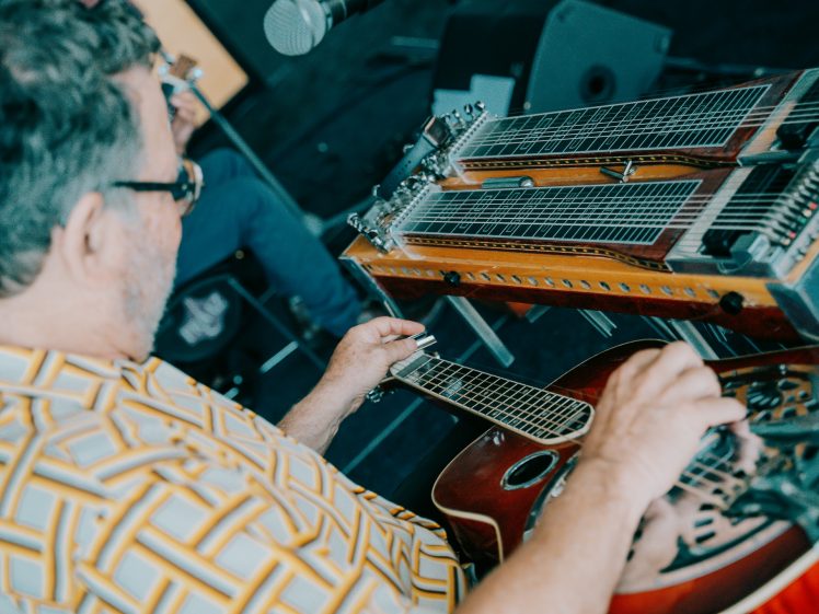Image of A man in a patterned shirt plays a resonator guitar with a steel slide next to a pedal steel guitar. The scene is set indoors with audio equipment visible in the background.