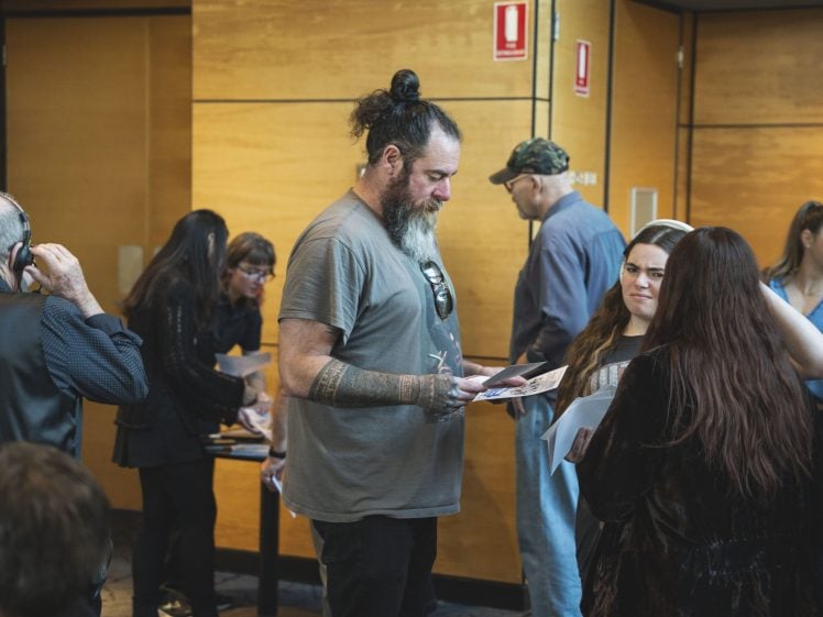 Image of A bearded man with a bun and tattoos reads a paper while standing in a group of people, some talking and some looking at documents, in a modern room with wooden walls.