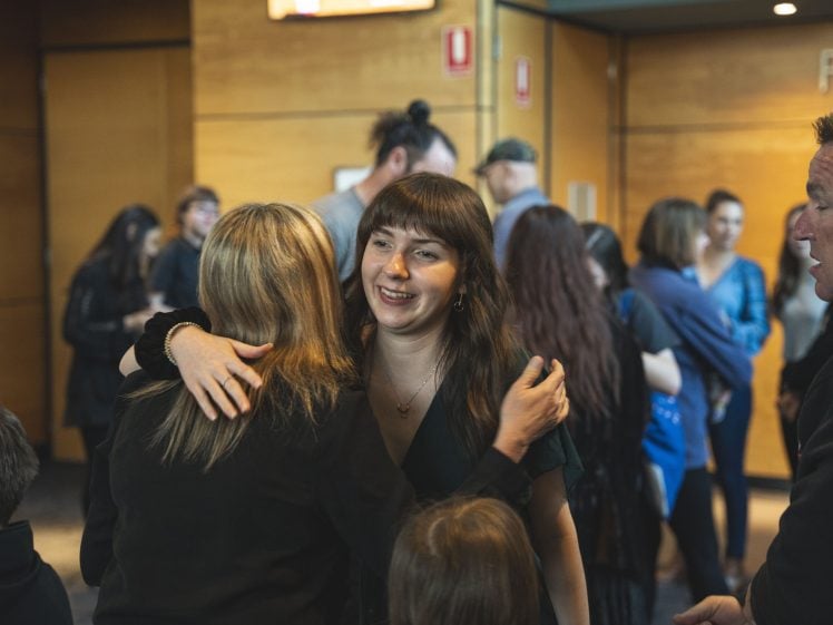 Image of A smiling woman embraces another person in a crowded indoor space, with several people conversing in the background. The atmosphere appears warm and friendly.