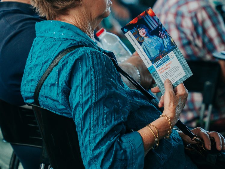 Image of An older woman in a textured teal blouse sits in a chair, holding a brochure and a water bottle, attentively listening during an indoor event with other seated attendees.