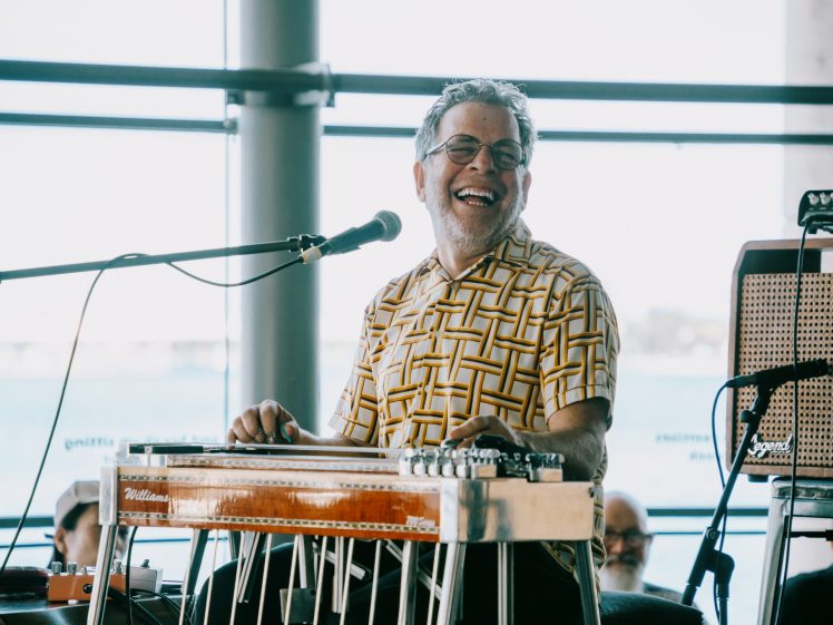 Image of A smiling man in glasses and a patterned shirt plays a pedal steel guitar and sings into a microphone onstage, with bright natural light and windows in the background.