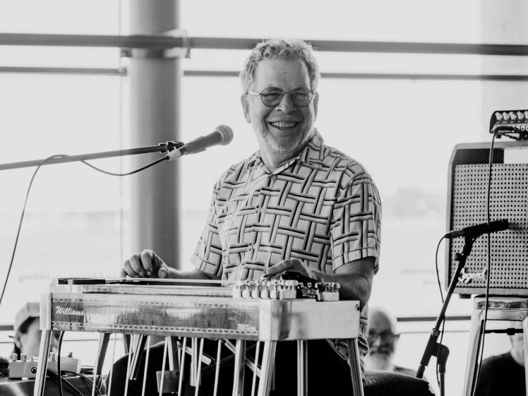 Image of A smiling man in glasses and a patterned shirt plays a pedal steel guitar and sings into a microphone. The scene is indoors with natural light streaming in through large windows.