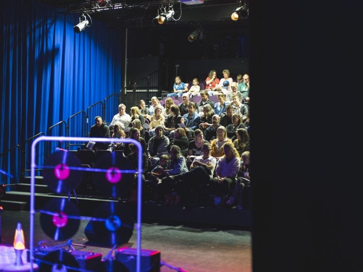 Image of An audience sits in a small theatre with blue curtains, waiting for a performance to begin. Colourful stage lights illuminate the space. Some stage equipment and props are visible in the foreground.
