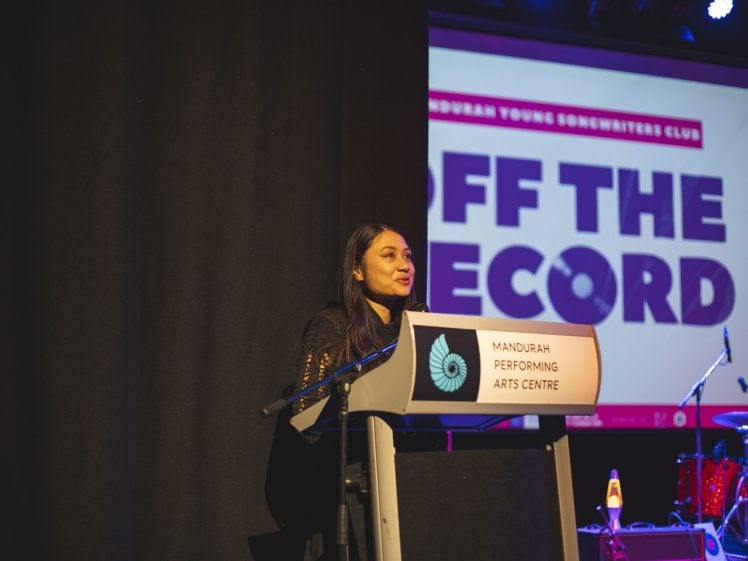 Image of A woman speaks at a lectern labelled Mandurah Performing Arts Centre with a colourful Off The Record event sign projected behind her on stage.