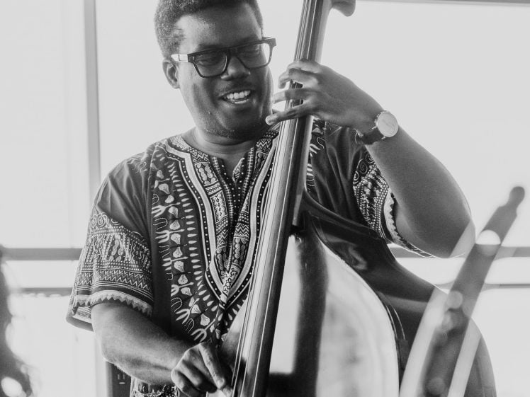 Image of A smiling musician wearing glasses and a patterned shirt plays a double bass indoors, with natural light streaming through large windows behind him. The image is in black and white.