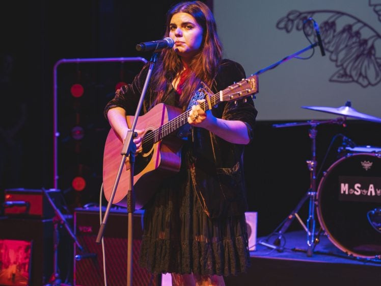 Image of A woman in a black dress and boots plays an acoustic guitar and sings into a microphone on stage, with drums and stage lights behind her.