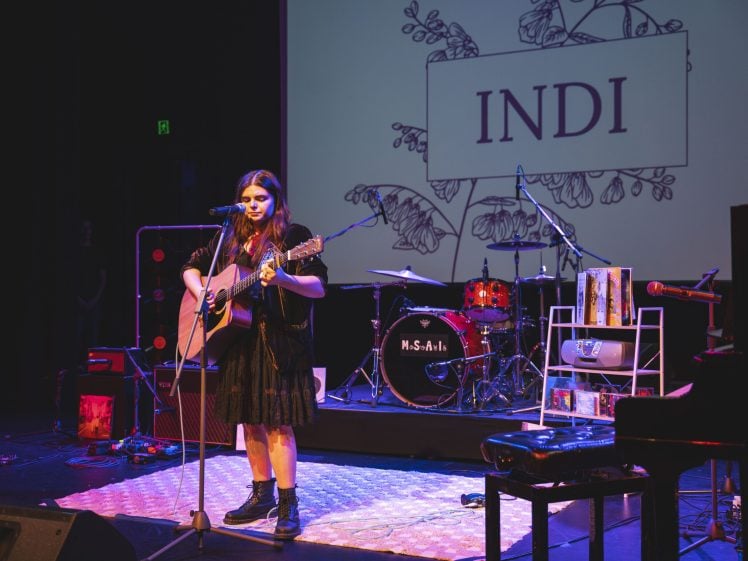 Image of A woman in a black dress plays an acoustic guitar and sings on stage. Behind her are drums, a keyboard, and a screen displaying INDI with floral art. The stage is lit with colourful lights.